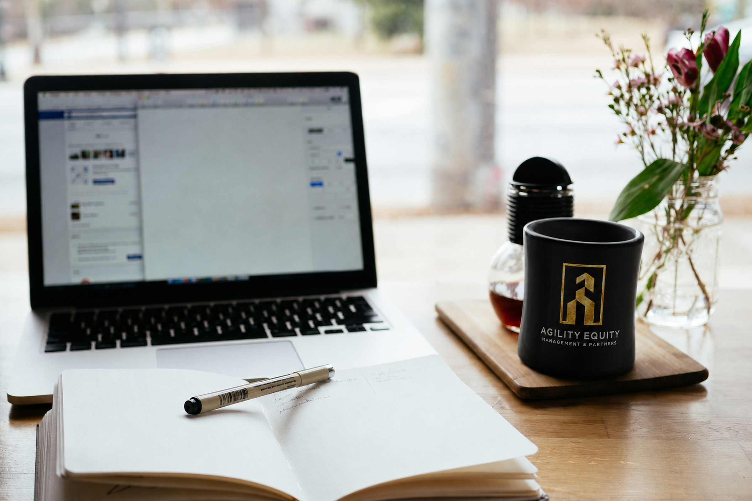 Desk with notebook, laptop, and a black Agility Equity logo mug